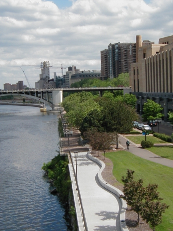 An urban river walkway with landscaping, office buildings, and bridge in background.