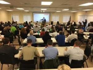 Photo of a group of men and women in a large room attending a presentation.