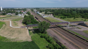Edge of a town with buildings near open fields. A bicycle and pedestrian bridge crosses multiple railroad tracks and a road. Several cyclists are riding on the path.