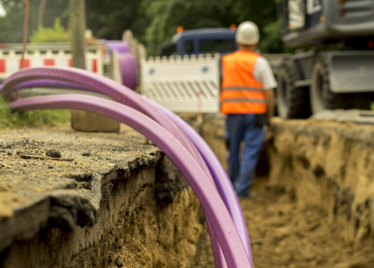 Fiber optic cables are being buried underground. A man working in the background stands in a dirt trench dug for the fiber, adjacent to a road where heavy equipment is parked.