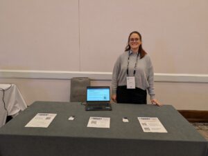 A woman stands behind a table. The table contains flyers with informational resources and a laptop showing newly released reports.